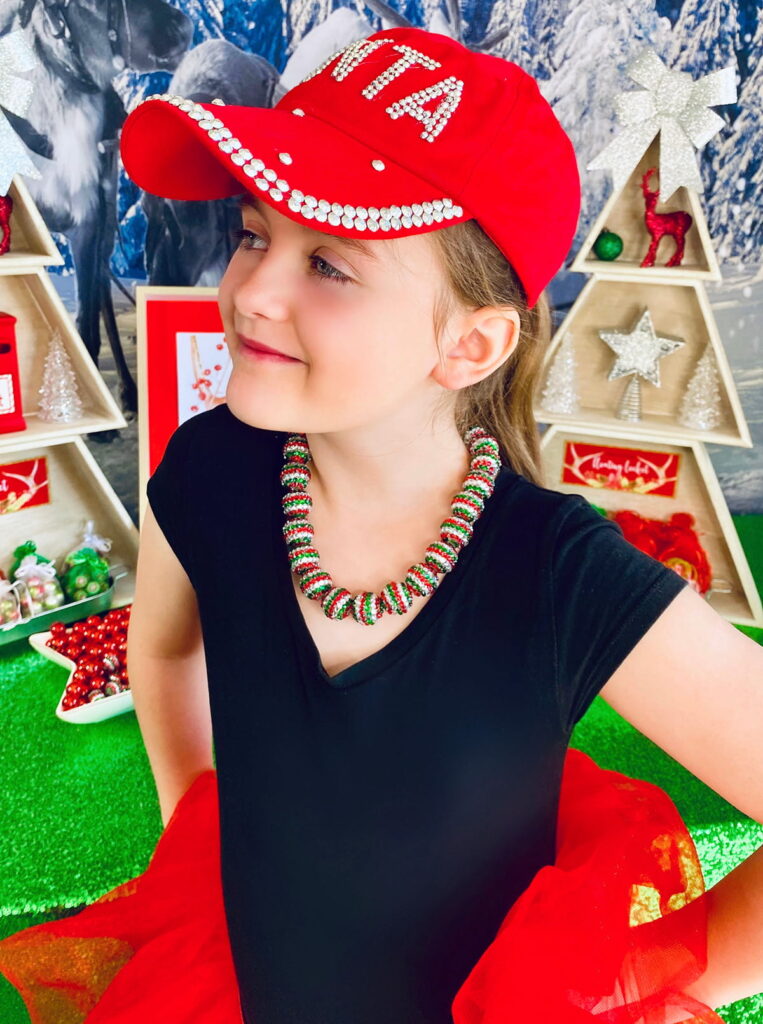 Girl wearing a Christmas bead necklace, looking off to the side in front of a kids’ Christmas jewelry-making party table.