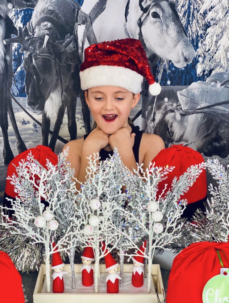 Girl in red sequin Santa hat smiling at a Christmas fairy garden table with mini elves, white and silver winter foliage picks, and red Santa-style supply bags.