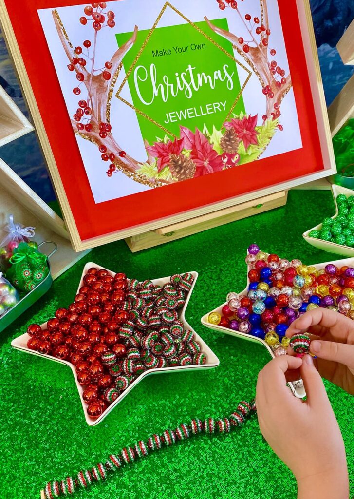 Young girl threading Christmas beads onto elastic at a party table, with star trays of colourful beads visible.