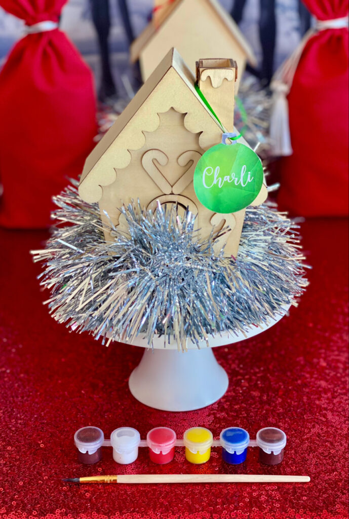 Undecorated wooden Christmas house on silver tinsel on a cake stand, with paint pots and a paintbrush in front, ready for decorating activity.