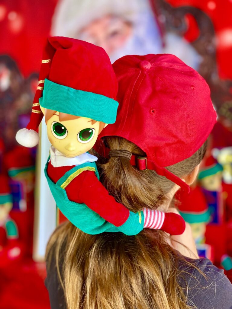 Plush elf perched on a child’s shoulders during an elf adoption party, showing the elf’s playful expression from behind the child.