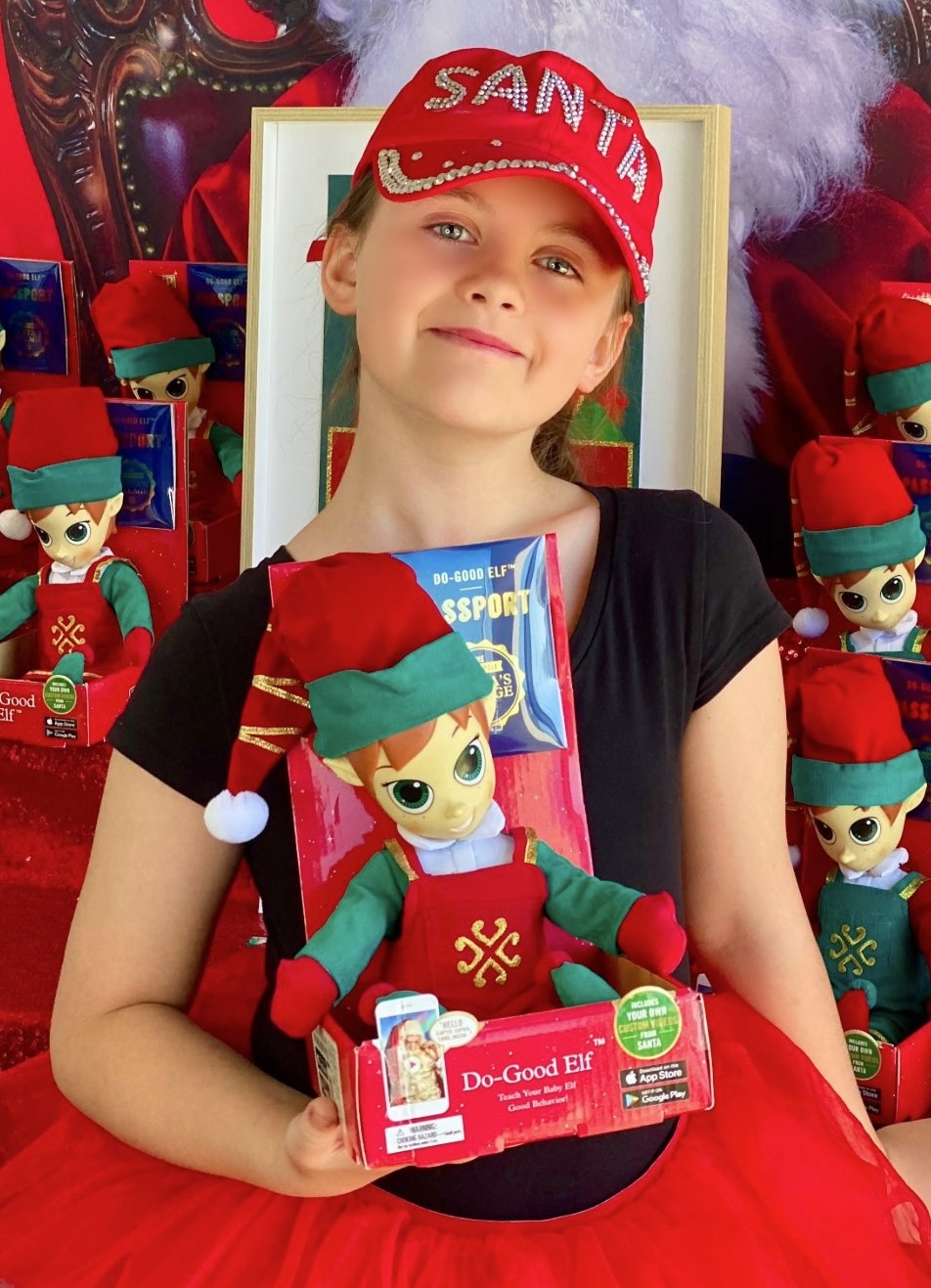 Child smiling with lips closed, holding the Portable North Pole elf she chose to adopt in front of the elf adoption party table.