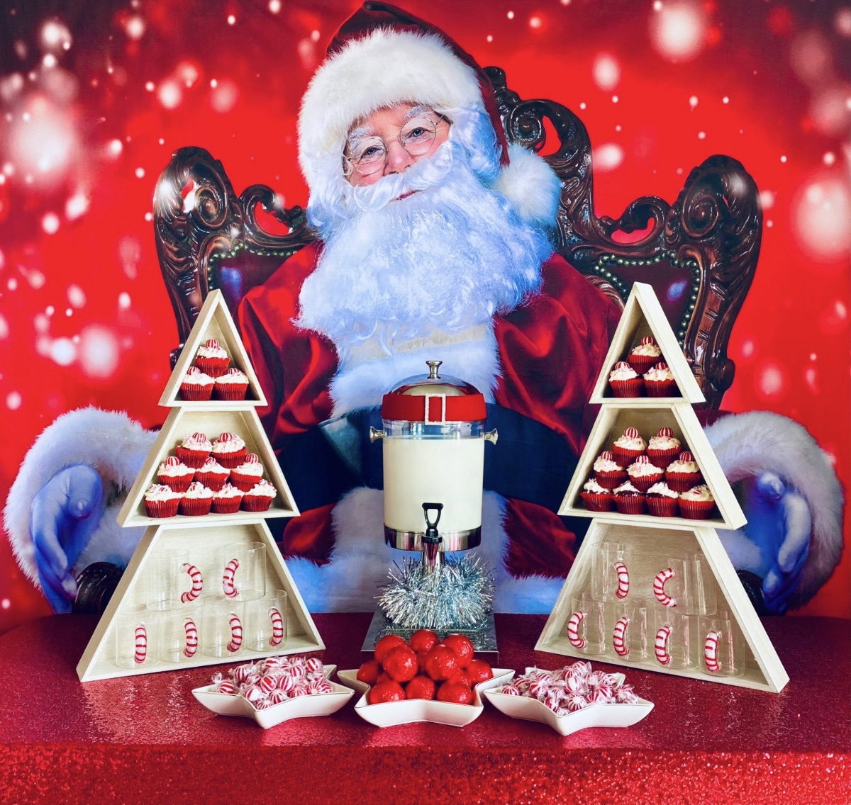 Christmas cocoa and cupcakes party table with Santa-themed backdrop, red sequin tablecloth, tree-shaped wooden shelves holding cupcakes and candy cane mugs, star platters with cocoa bombs and peppermints, and a milk dispenser.