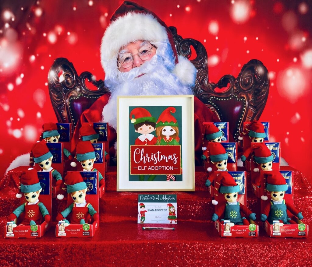 Elf adoption party table with Santa backdrop, red sequin tablecloth, plush elves in different sizes, Elf Adoption Party sign, and adoption certificates on display.