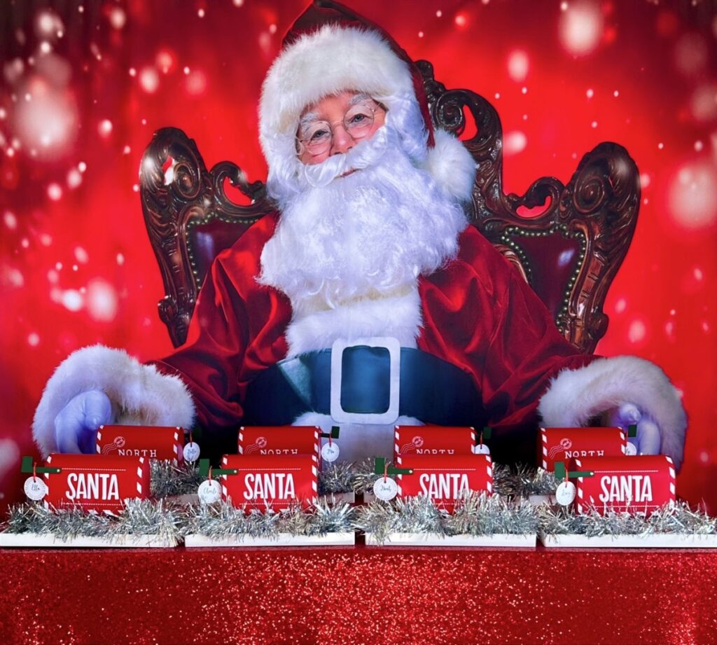 Christmas sand art party table with Santa backdrop, red sequin tablecloth, white trays trimmed with silver tinsel, and red Santa mailbox containers holding sand art supplies.