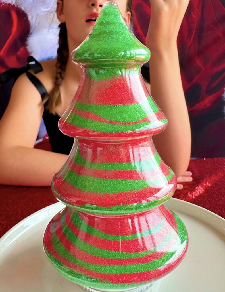 Completed Christmas tree sand art filled with red and green sand, with a girl blurred in the background at a kids Christmas sand art party.