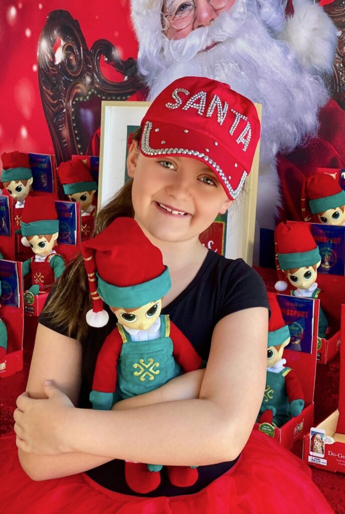 Young girl in a Santa cap holding her plush elf to her chest while standing in front of an elf adoption party table.