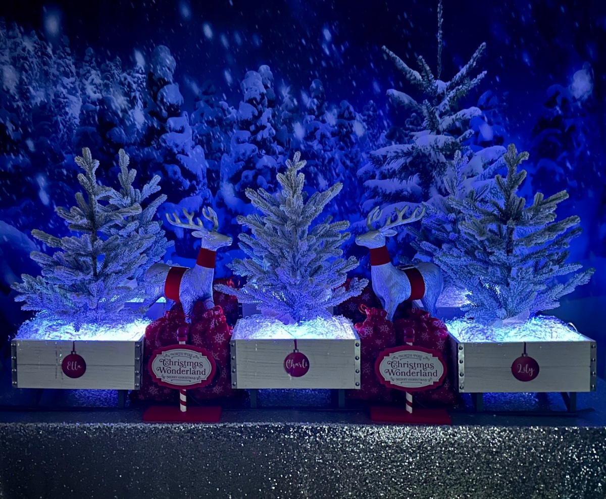 Glowing mini Christmas tree decorating party table with a snowy tree backdrop, silver sequin tablecloth, sleigh crates with iridescent shred, snow-tipped tabletop trees, red activity supply bags, and mini Christmas signs.