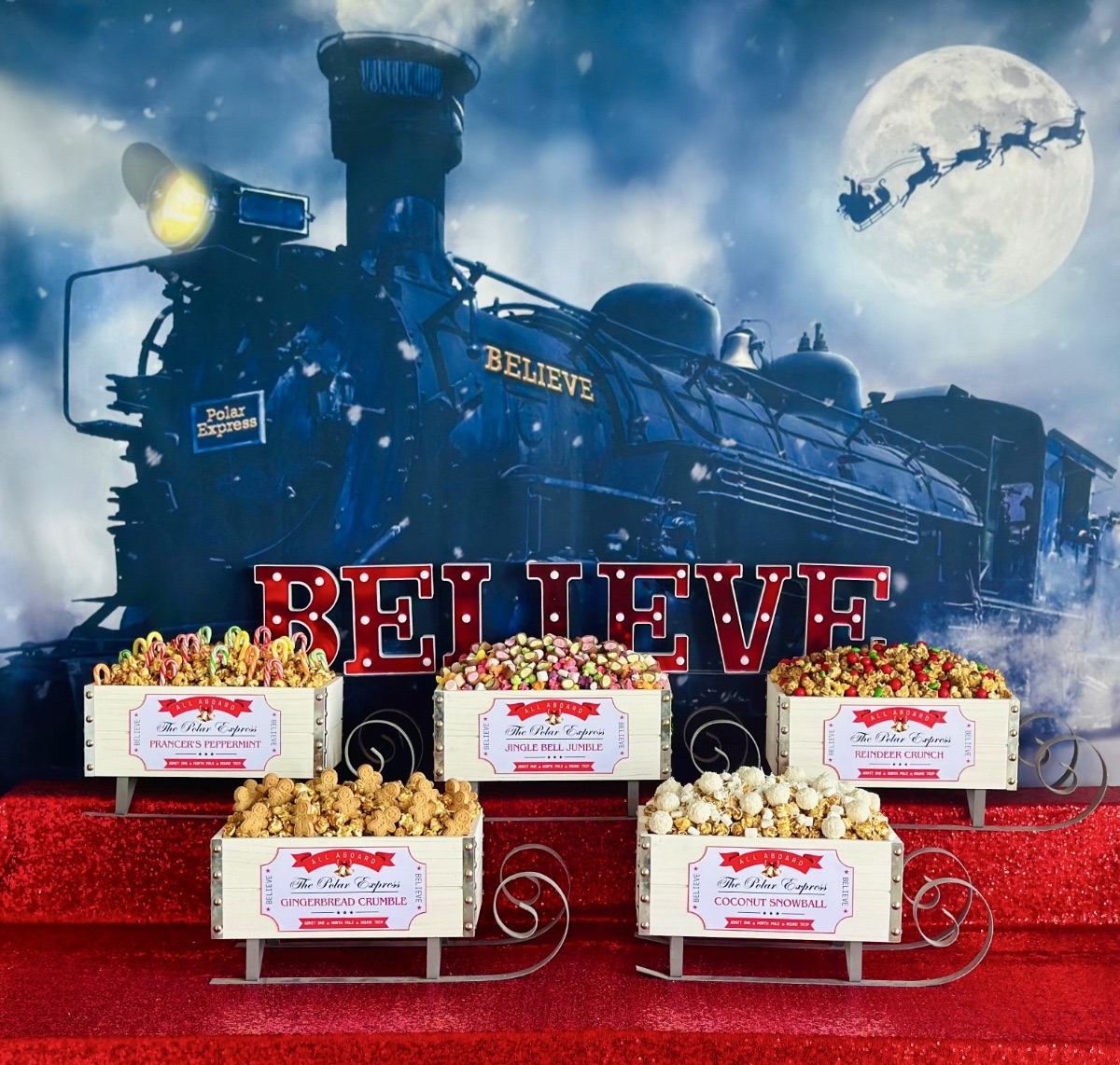 Polar Express Christmas popcorn party table with red sequin tablecloth, train backdrop, staggered white sleigh crates filled with flavoured popcorn, ticket labels, and red “Believe” marquee letters.