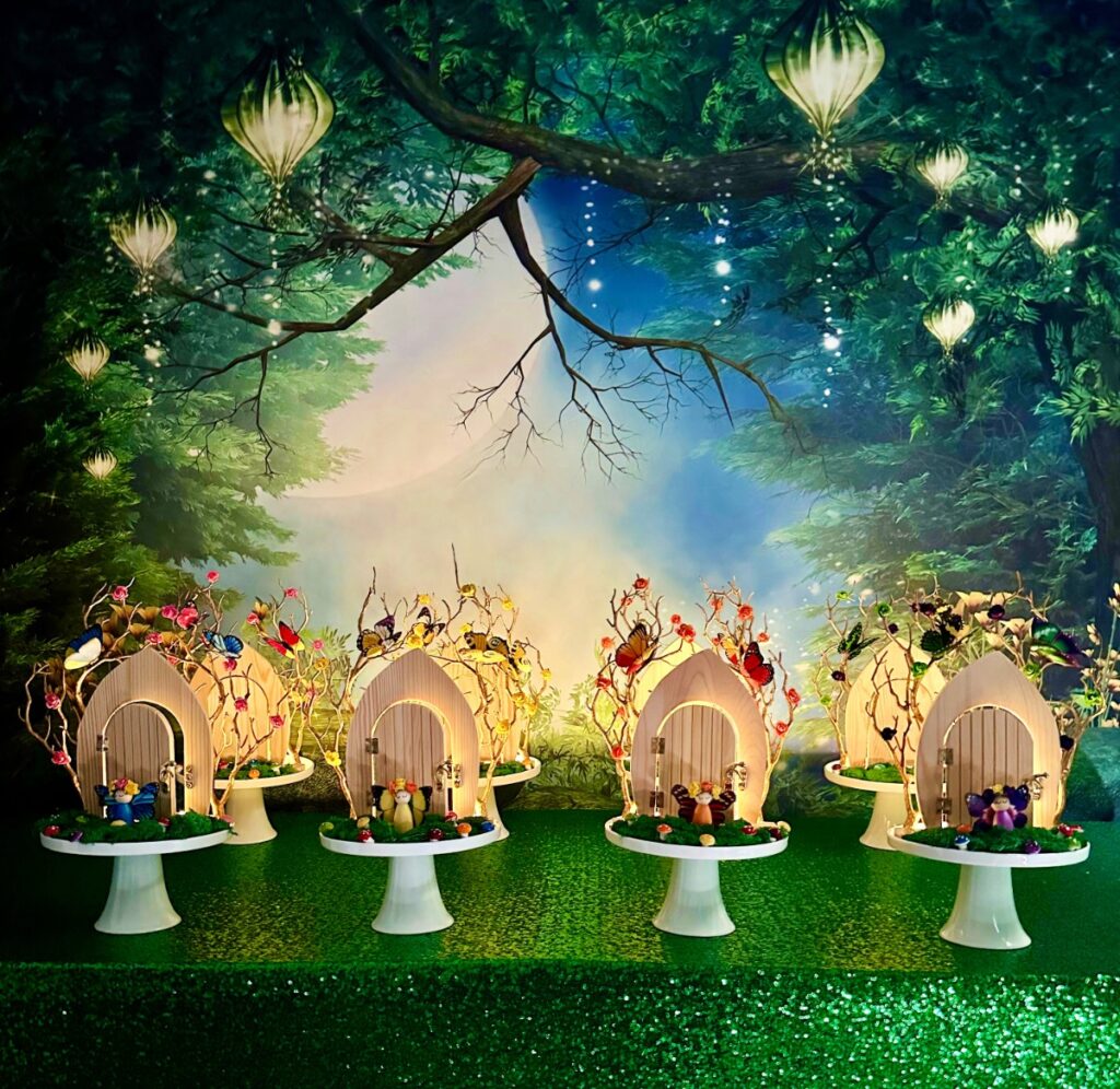 DIY fairy door party table with fairy garden backdrop, green sequin tablecloth and wooden fairy doors lit up with fairy lights ready to decorate.