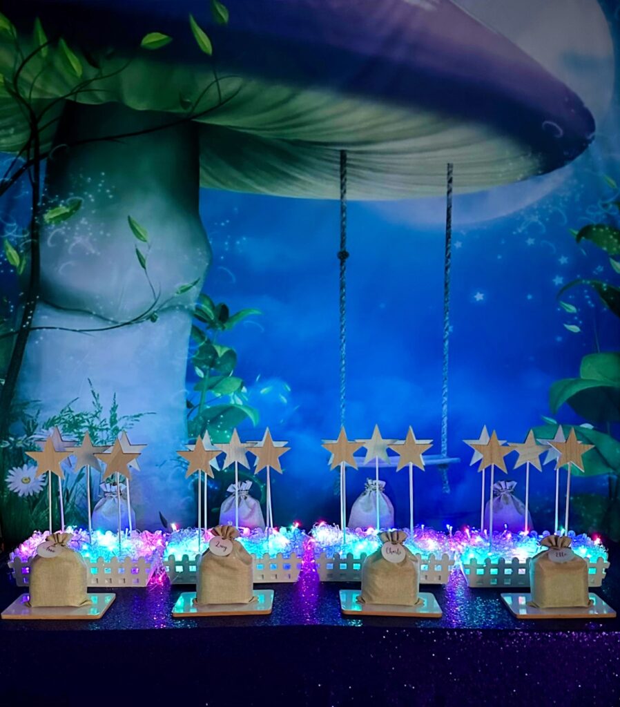 DIY fairy wand party table with purple mushroom swing backdrop, purple sequin tablecloth, wooden star wands in crates, fairy lights and hessian supply bags.