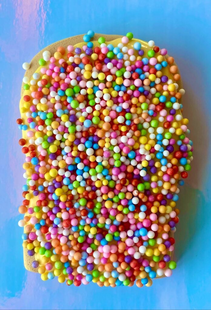 Close up of fairy bread slime on a board with foam balls that resemble hundreds and thousands sprinkles on butter slime and a clay bread shape.