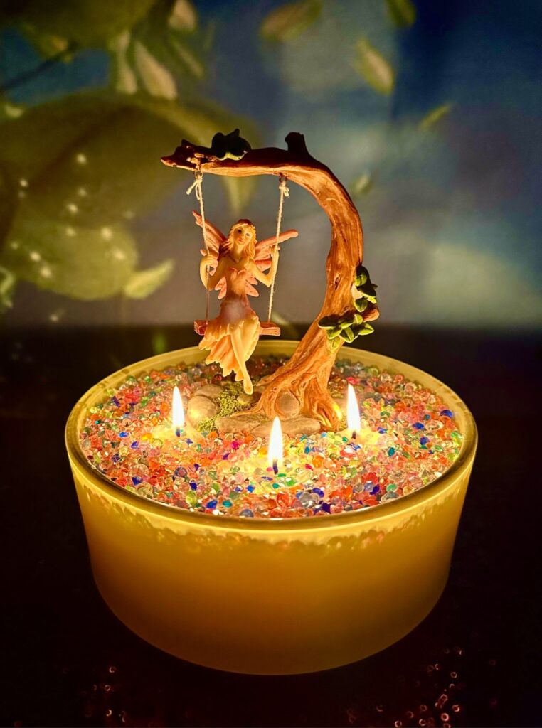 Fairy candle decorating party activity with yellow candle, fairy on a swing figurine in centre and coloured glass on top of candle.