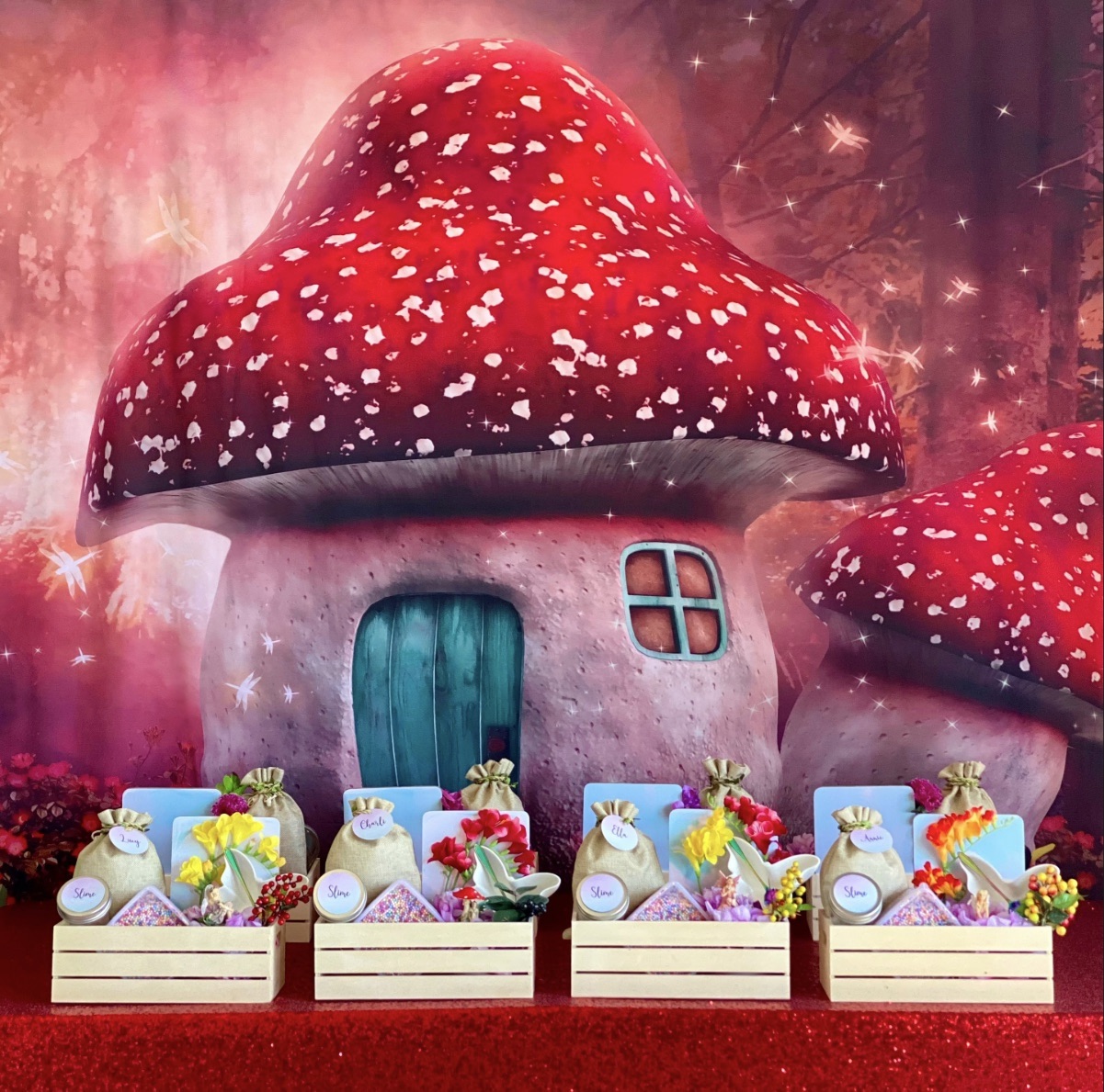 Fairy slime party table with mushroom backdrop, red sequin tablecloth, wooden crates of slime supplies, butterfly dish, faux flowers and clear take-home jars.