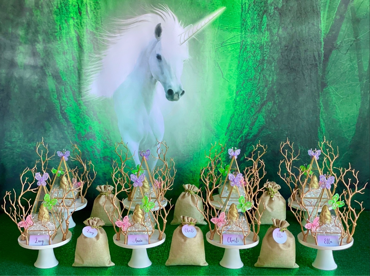 Kids unicorn slime party table with white unicorn backdrop, cake stands holding terrariums with gold horns and hessian slime supplies bags.