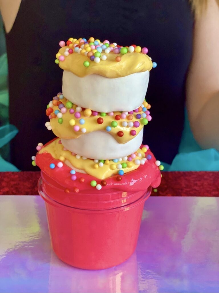 Stacked fairy bread slime in a jar, with a pink slime base, clay rounds on top, butter slime topping, and foam sprinkles like hundreds and thousands.