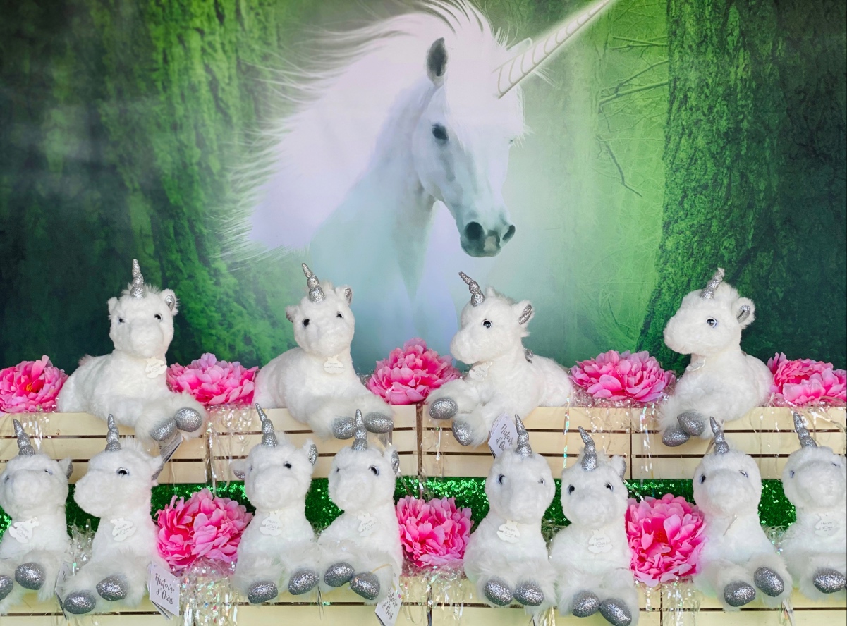 Close-up of a Unicorn Adoption Party table with a unicorn backdrop and staggered wooden crates filled with plush white unicorns, pink flowers, and iridescent shred.