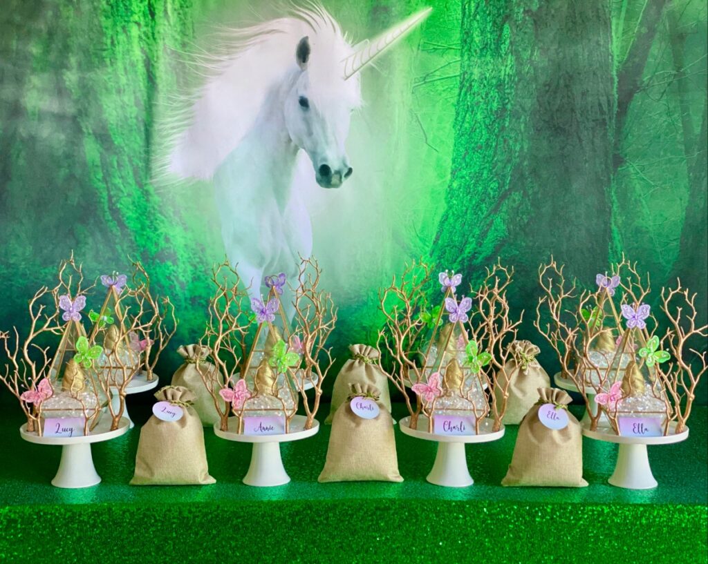 Unicorn horn slime party table with unicorn backdrop, green sequin tablecloth, white cake stands and gold terrariums filled with iridescent shred and gold unicorn horns.