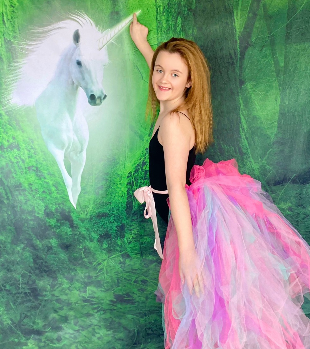 Young girl in a pastel tutu smiling and touching a unicorn horn on a unicorn party backdrop during a kids’ Unicorn Adoption Party.