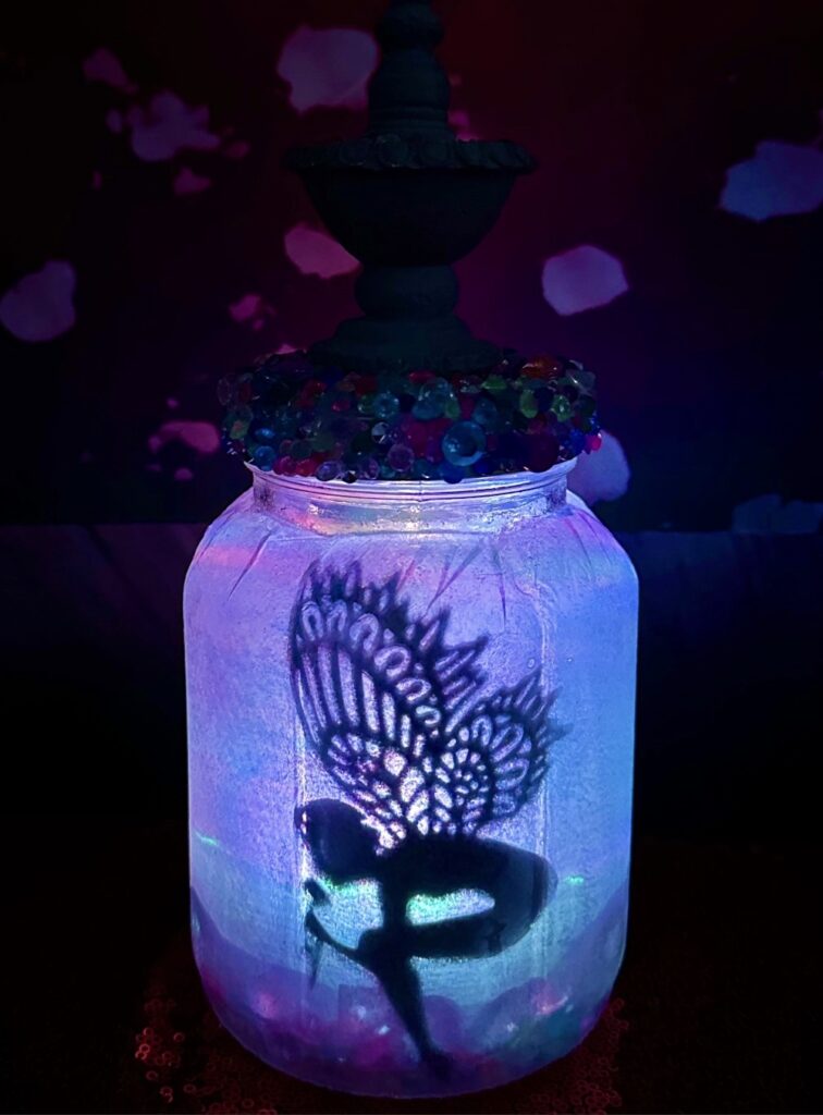 DIY fairy lantern made from a mason jar with a glowing blue LED light, featuring a fairy silhouette with beautiful wings bending to pick a flower.