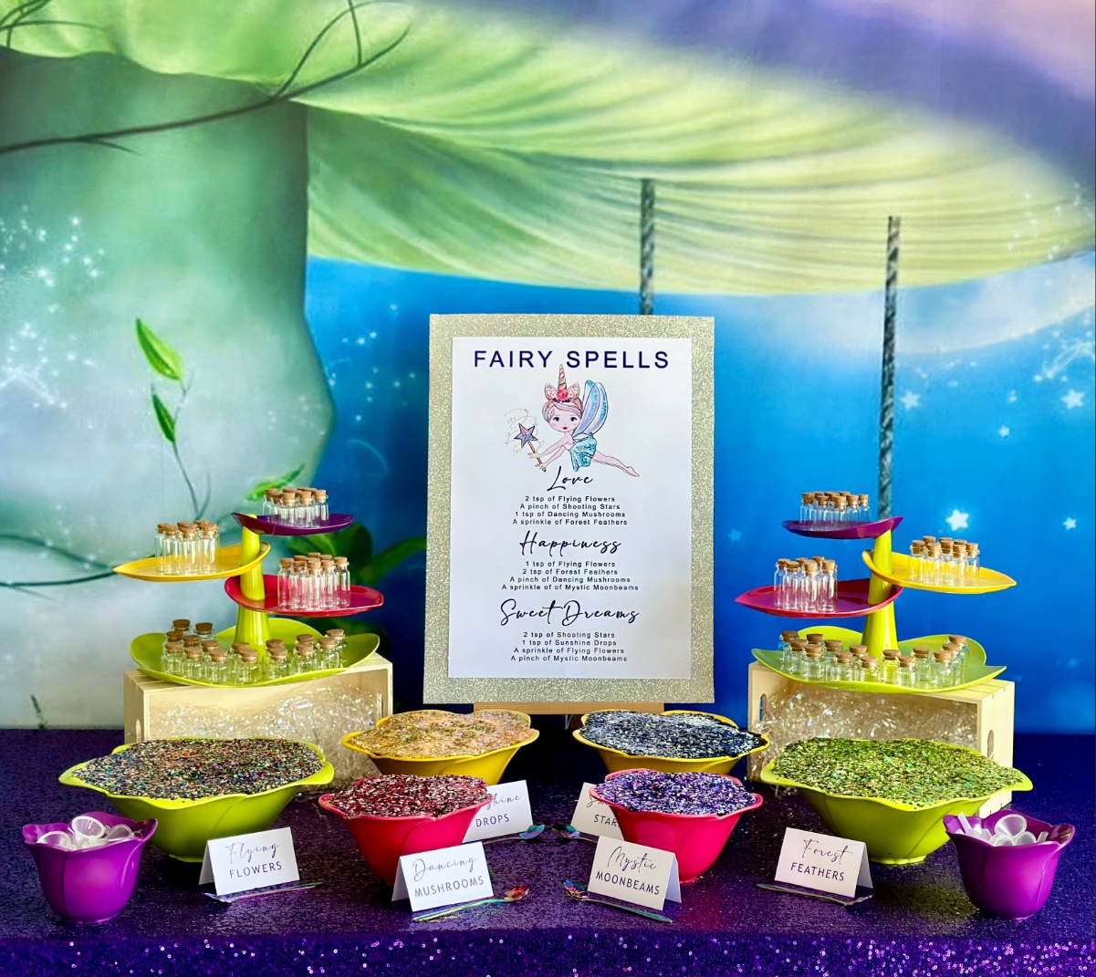 DIY fairy potion party table with a purple mushroom swing backdrop, purple sequin tablecloth, leaf platters holding mini glass jars, flower bowls filled with glitter for a kids fairy party.