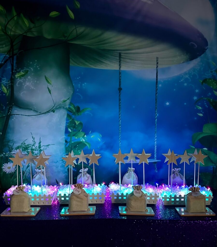 DIY fairy wand party setup with purple sequin tablecloth, mushroom swing backdrop, glowing fairy lights, star wands in white crate displays and hessian bags with decorating supplies.