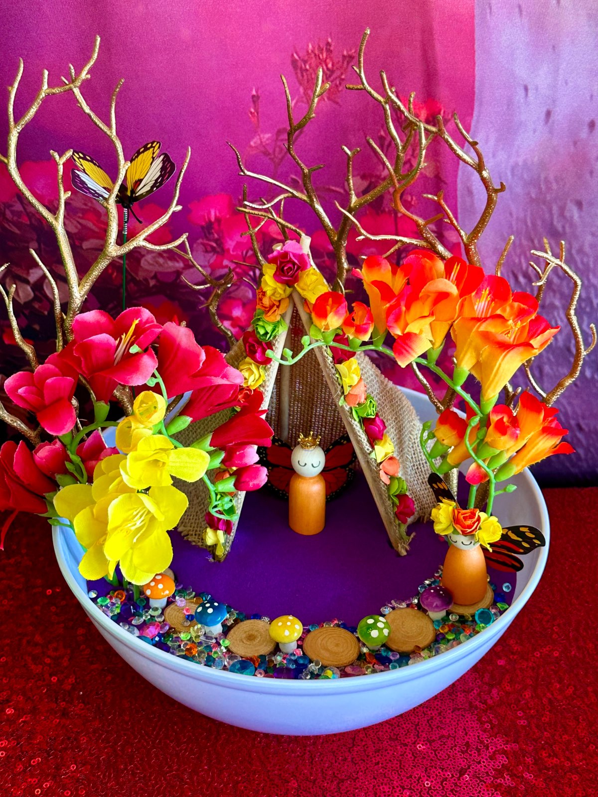 Close-up of fairy garden with purple sand, colourful faux flowers, gold twigs, flower-trimmed hessian teepee, orange wooden fairies, wooden path and crystals.