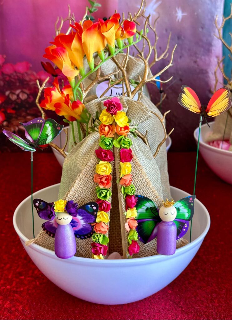 Fairy garden party activity kit in a white bowl with mini purple wooden fairies, hessian bags of supplies, faux flowers and a flower-trimmed hessian teepee.