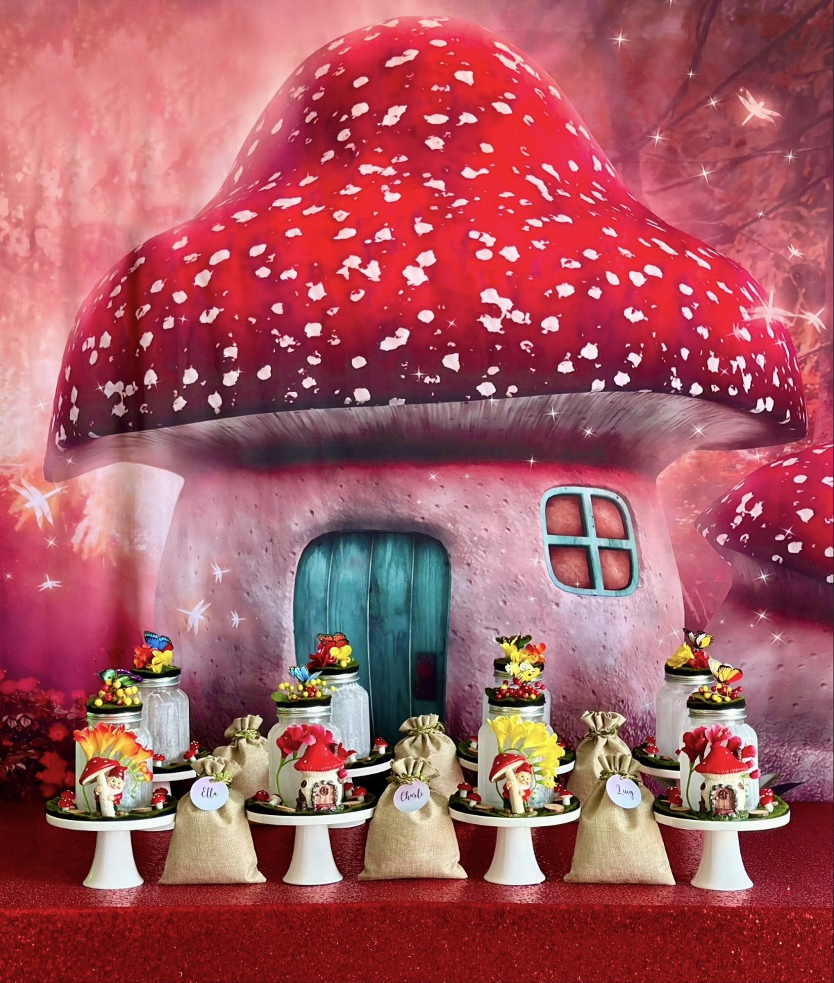 DIY fairy lantern party table, with a fairy mushroom house backdrop, red sequin tablecloth, mason jars on cake stands, fairy decorations, and hessian bags filled with supplies.