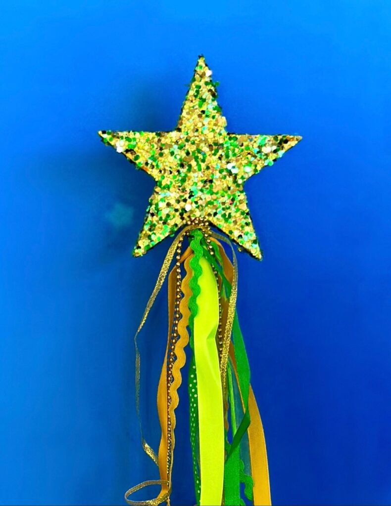 Close-up of green and gold glitter fairy wand with yellow and green ribbons and decorative trims hanging from the handle.
