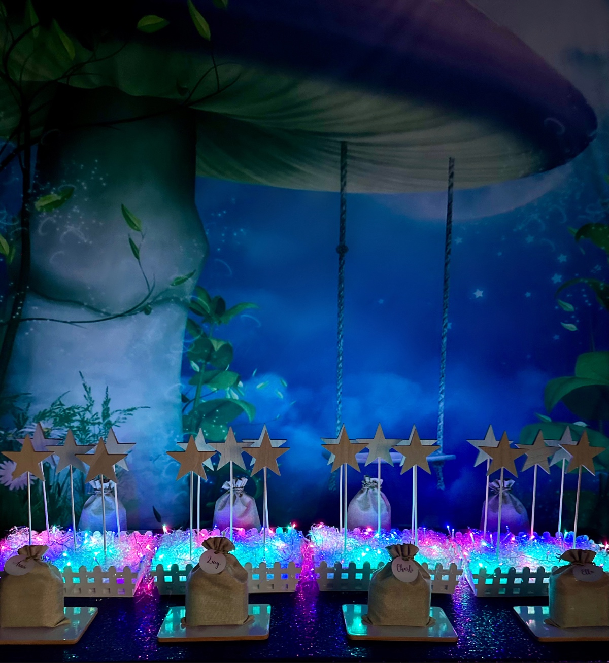 DIY fairy wand party setup with purple sequin tablecloth, mushroom swing backdrop, hessian bags and glowing fairy lights illuminating star wands in white crate displays.