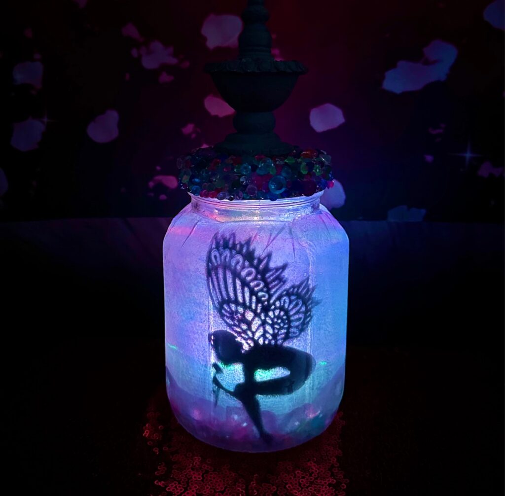 Glowing purple fairy jar lantern DIY featuring a fairy silhouette with beautiful wings bending to smell a flower inside the jar.