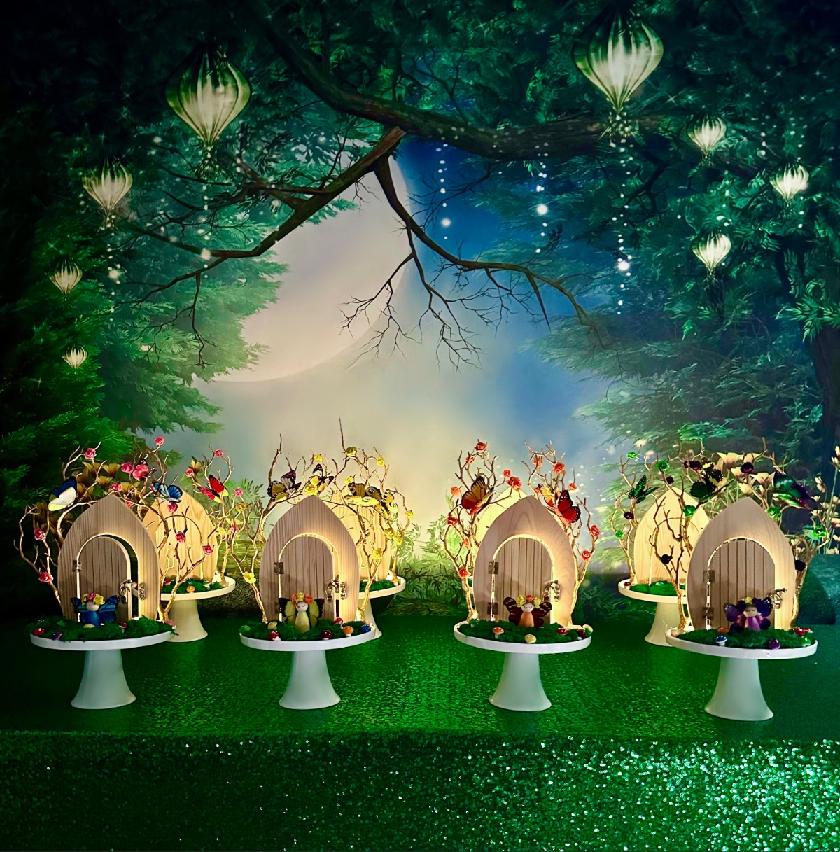 DIY fairy door party setup featuring glowing fairy doors on moss-covered stands, fairy figurines, mushrooms, green sequin tablecloth, and a fairy garden backdrop.