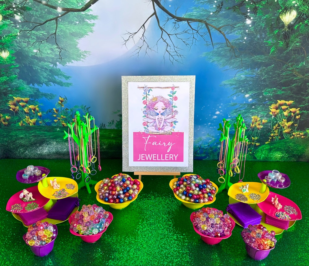 DIY fairy jewelry party table with a fairy garden backdrop, green sequin tablecloth, fairy jewelry sign, leaf-shaped platters with charms, floating lockets hanging from jewelry trees, and flower-shaped bowls filled with beads.