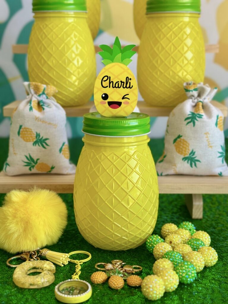 DIY pineapple jewelry kids party activity supplies including a kawaii pineapple jar, bubblegum beads, pineapple charms, a floating locket keychain, letter charm, and yellow pom poms.