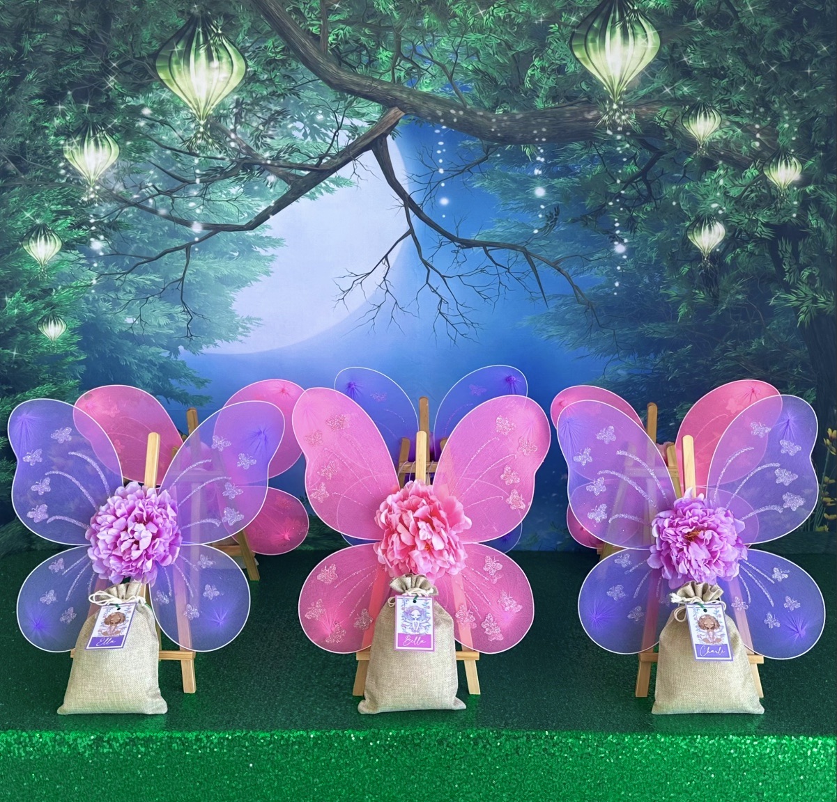 DIY fairy wings party table with a green sequin tablecloth, fairy garden backdrop, and pink and purple wings on easels with hessian bags of decorating supplies.