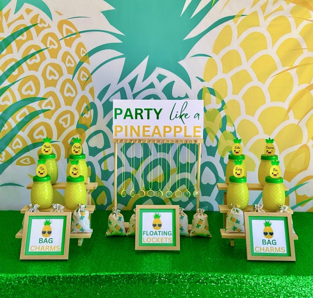Kids DIY pineapple party table setup with pineapple backdrop, green sequin tablecloth, floating lockets hanging on a jewelry stand, and tiered shelves with pineapple jars and supply bags.