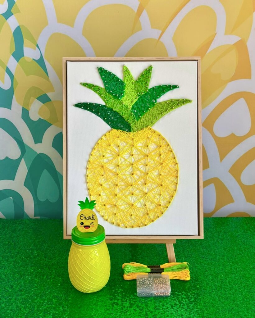 Kids pineapple string art party featuring a completed pineapple string art canvas on an easel with crafting supplies and a pineapple jar in front.