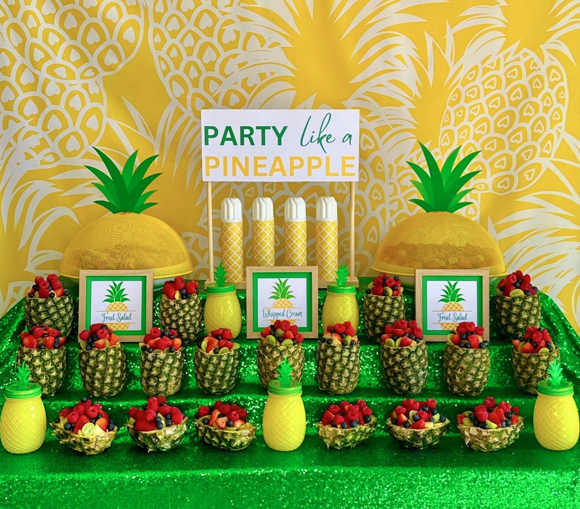 Pineapple fruit salad station for a kids party with berry fruit salad served in pineapples, a Party Like a Pineapple sign, and tropical table decorations.