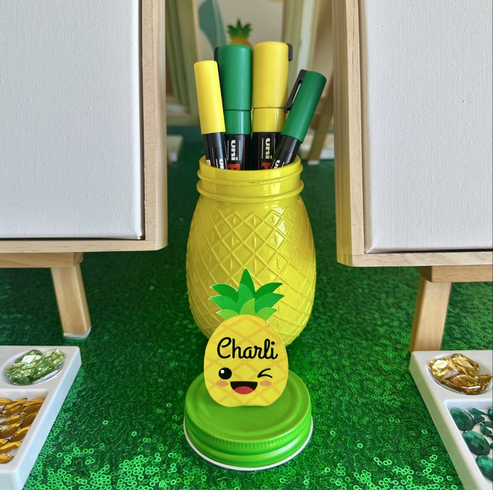 Close-up of pineapple painting party supplies with Posca paint pens in a pineapple jar and a kawaii pineapple name tag for a kids party activity.