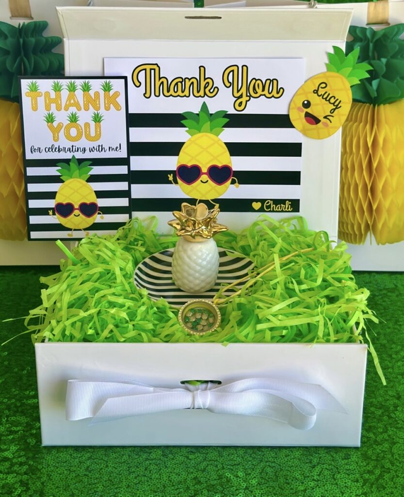 Kids pineapple party favour featuring a pineapple trinket dish and a yellow floating locket filled with pineapple charms, presented in a white gift box with a thank you card and name tag.