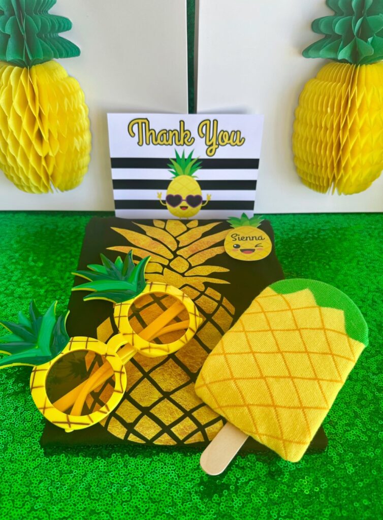 Kids pineapple party favour featuring pineapple popsicle socks, pineapple novelty glasses, a pineapple T-shirt, and a pineapple-themed thank you card displayed with decorated gift bags.