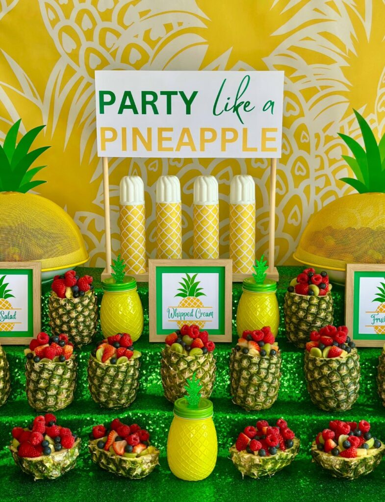 Kids pineapple party fruit salad station with hollowed-out pineapples filled with fruit salad, a pineapple backdrop, green sequin tablecloth, and tropical decorations.
