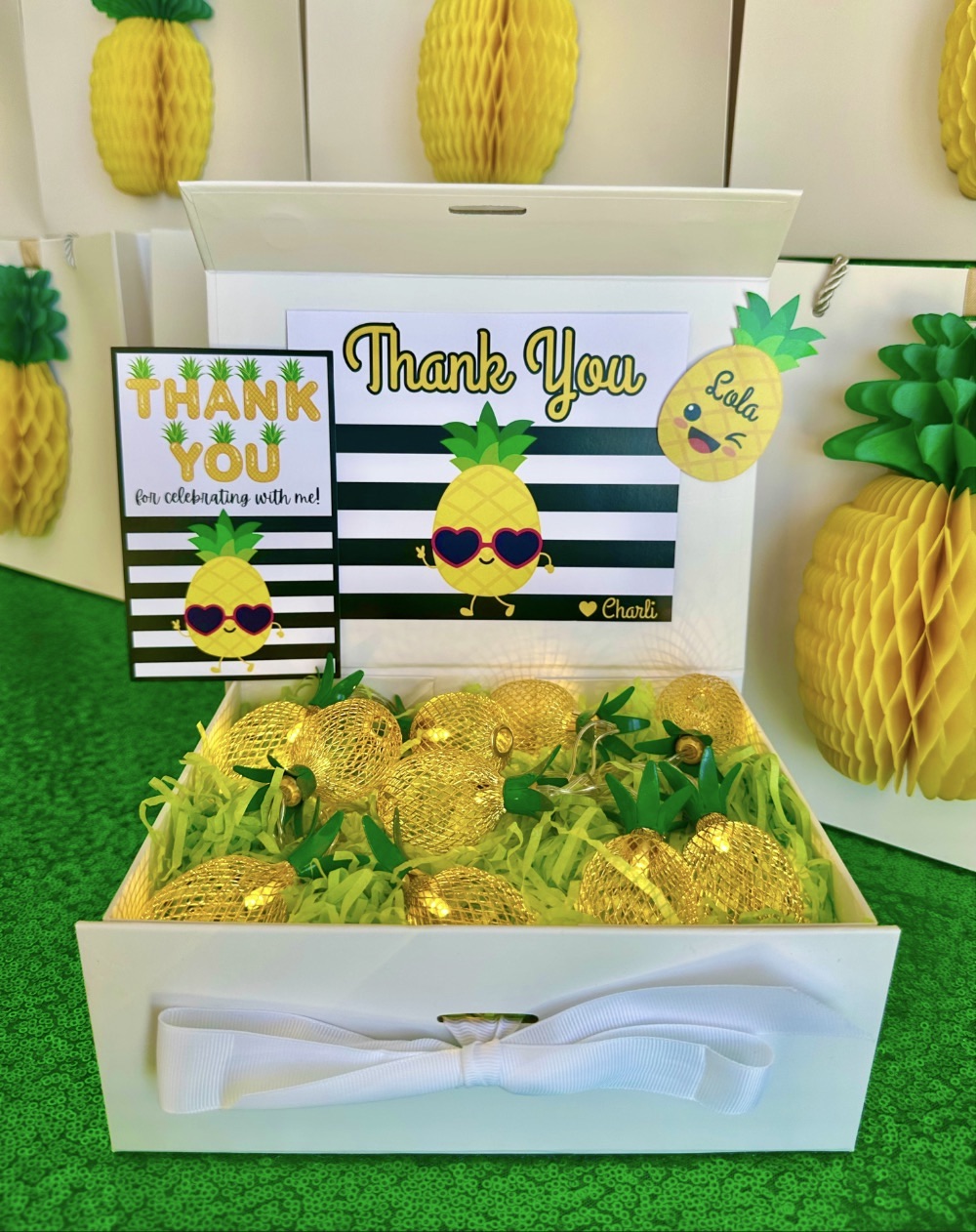 Pineapple-shaped string lights displayed in a gift box with pineapple-themed thank you cards and name tag, with gift bags in the background.