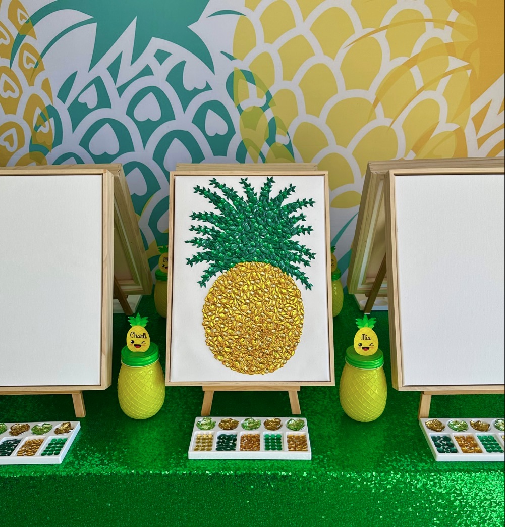 Kids pineapple painting party table with canvases on easels, a gem-decorated pineapple artwork, pineapple supply jars, and acrylic gem palettes.