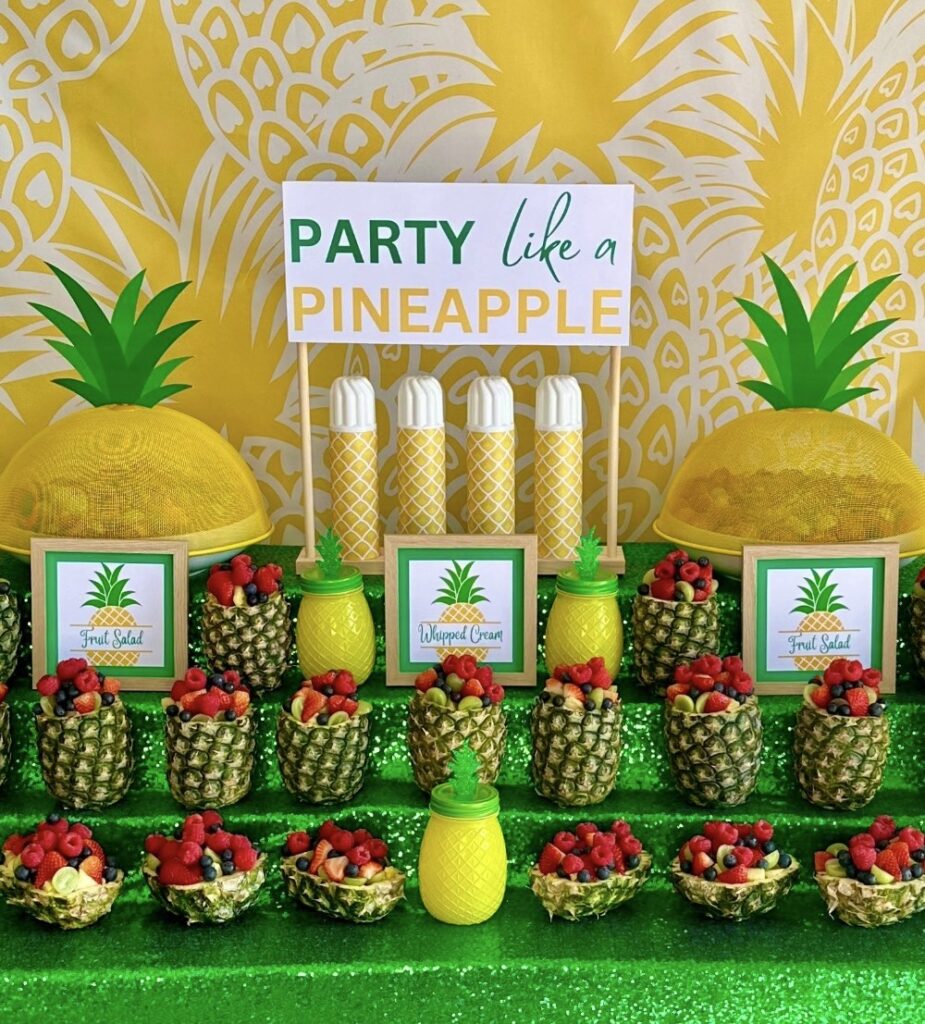 Kids pineapple party fruit salad station featuring hollowed-out pineapples filled with fresh fruit, tropical party decorations, a pineapple-themed backdrop, and a green sequin tablecloth.