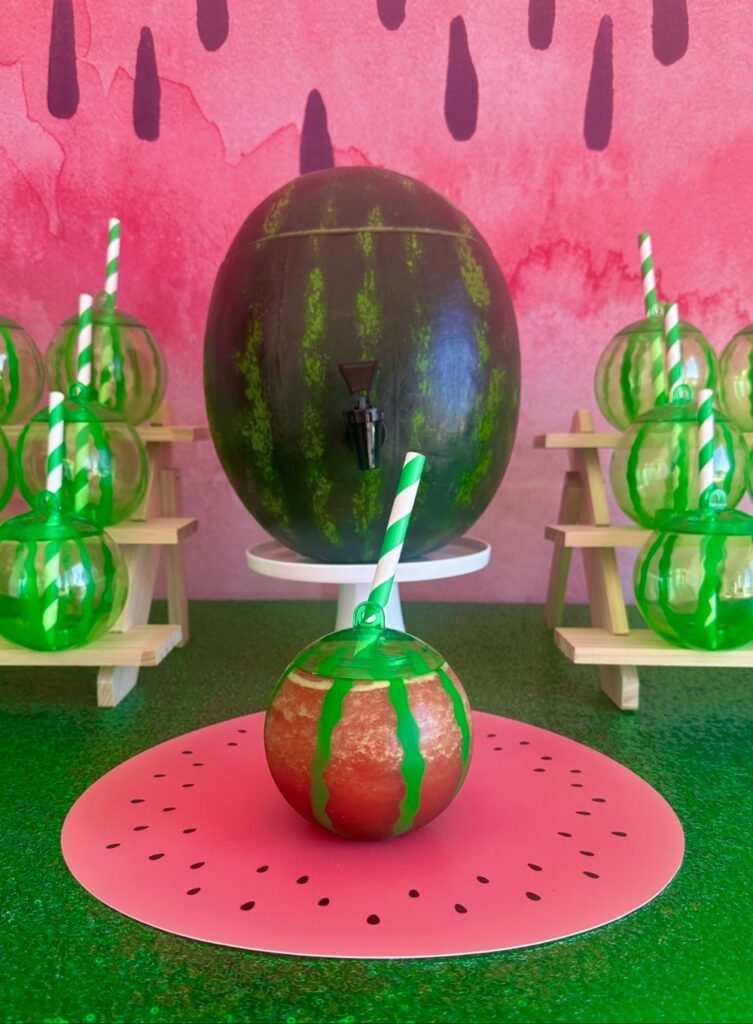 Kids watermelon party drink station with a watermelon keg, watermelon cups and straws on a green sequin tablecloth.