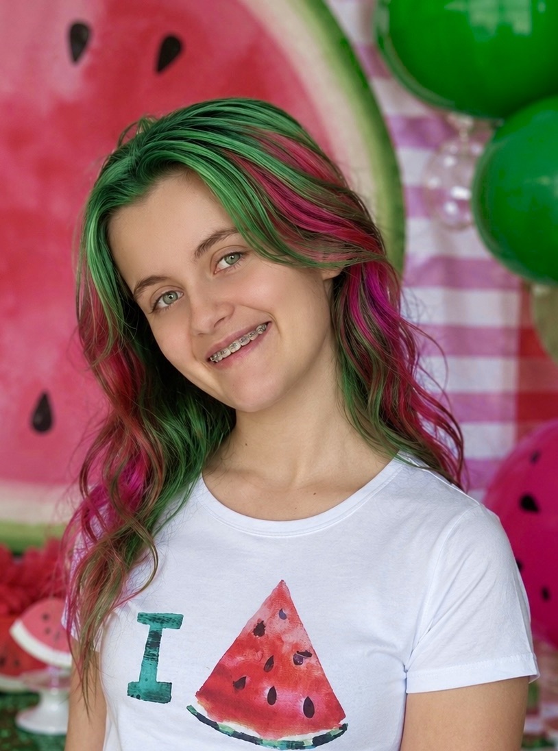 Smiling girl with pink and green hair wearing a watermelon-themed shirt in front of a watermelon party backdrop.