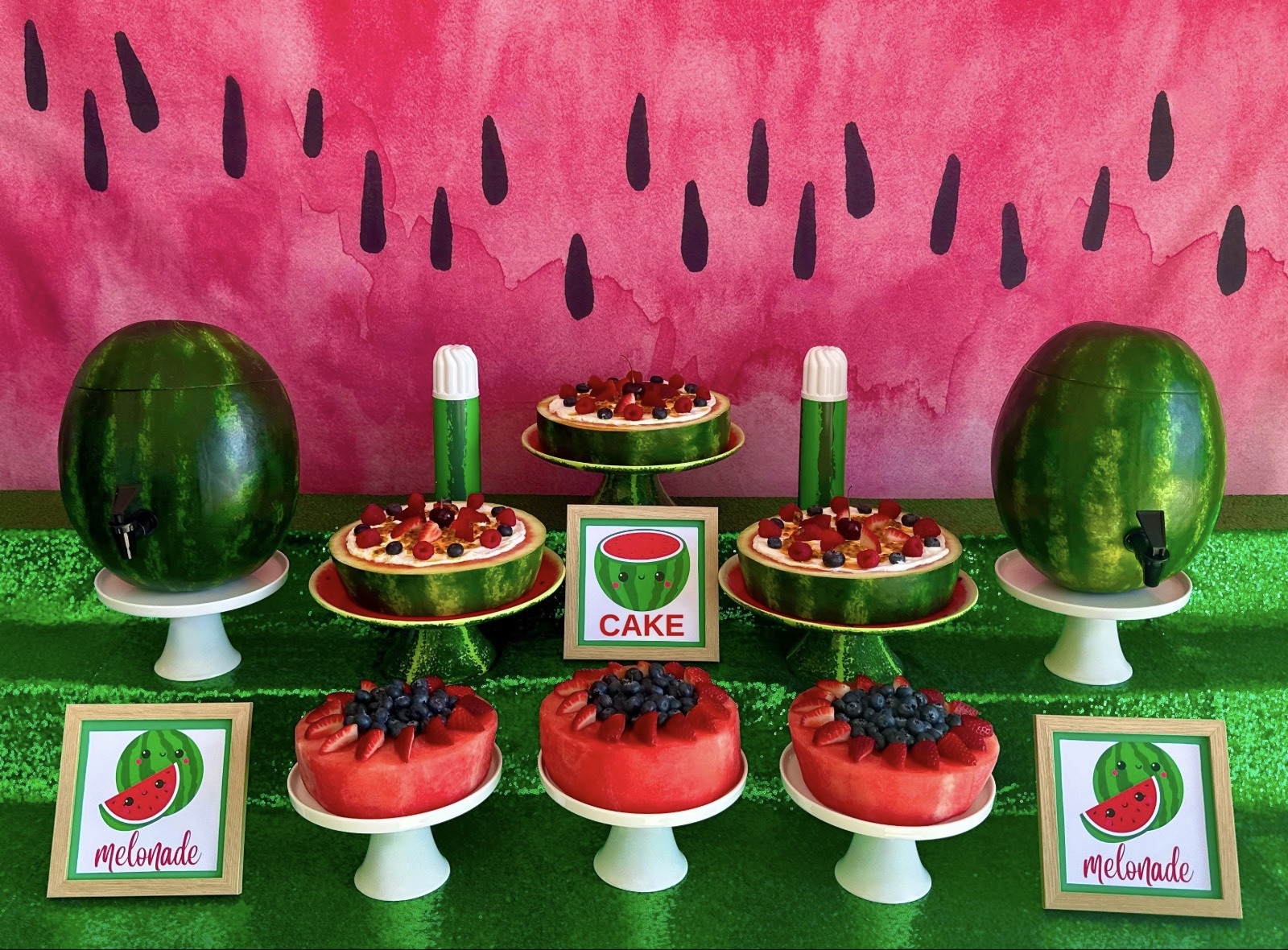 Fresh watermelon cake party table with berry-topped cakes, melonade dispensers, green sequin tablecloth and pink watermelon backdrop.