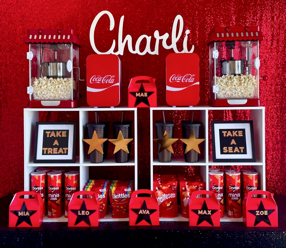 Kids’ movie party snack bar with popcorn machines, mini drinks fridges, candy, chips, personalised tumblers and red snack boxes displayed across the front.